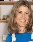 Woman in a blue shirt and white apron waving in front of shelves with various items.