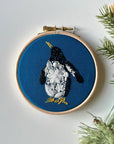 Embroidery hoop with a penguin design on blue fabric, surrounded by greenery.
