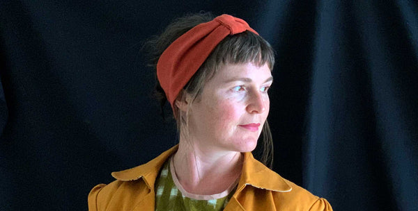 an image of a white woman wearing a rust coloured headband the slightly covers her ears. The image shows the side of the woman face looking off camera. She is wearing a mustard yellow jacket and a green and white check shirt. it has a black background