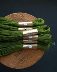 Four skeins of olive green embroidery floss with gold paper bands, arranged on a round wooden board against a dark background.