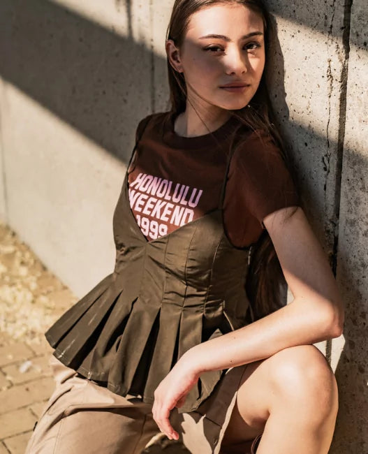 Woman wearing a brown t-shirt with text and a green pleated top, sitting against a concrete wall.