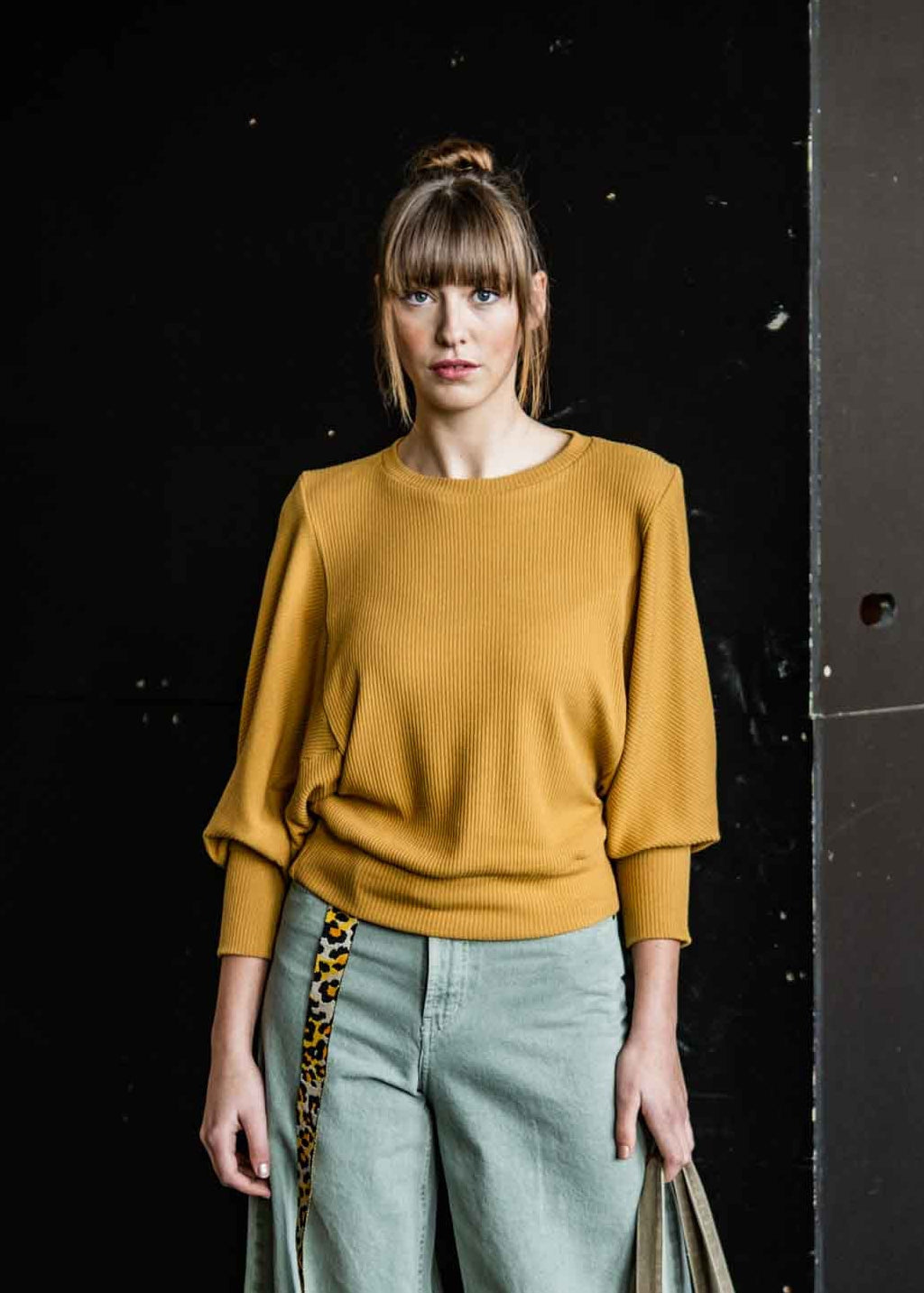 Woman wearing a mustard yellow sweater and light blue pants, holding a beige tote bag against a dark background.