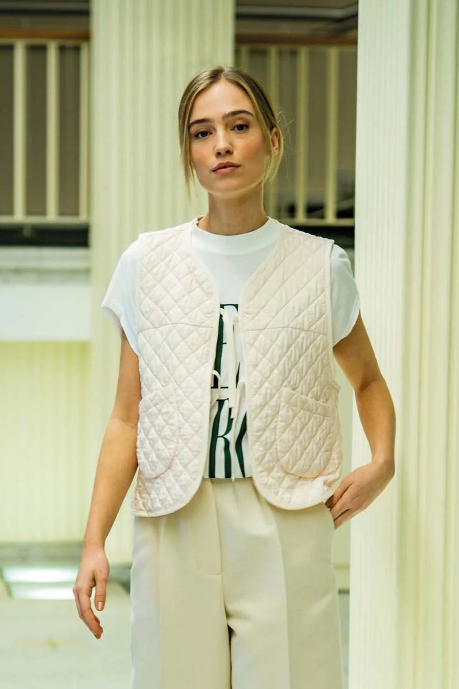 Woman wearing a light-colored vest over a white shirt with a patterned tie, standing in a modern indoor setting.