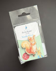 A Peter Rabbit Needle Mint pin in its original clear plastic packaging, featuring an illustration of a squirrel and a rabbit, set against a dark gray background.