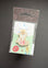 A Peter Rabbit enamel pin in its original packaging, featuring a rabbit holding a pink teacup on a light green backing card with a brown hanging tab, set against a dark gray background.