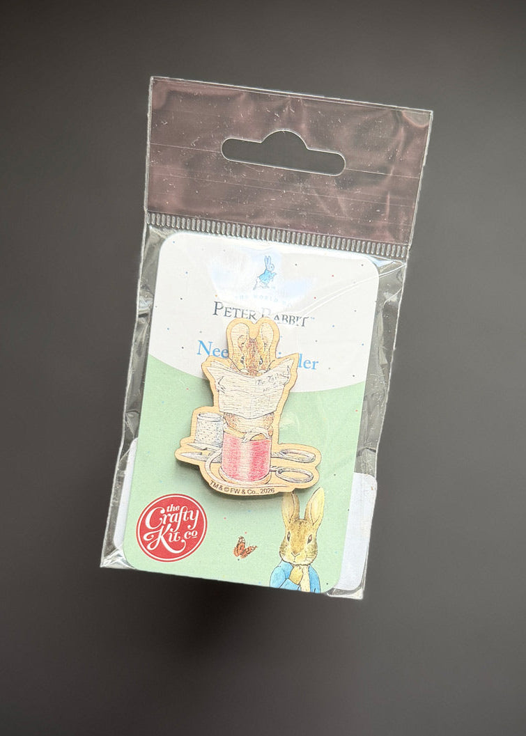 A Peter Rabbit enamel pin in its original packaging, featuring a rabbit holding a pink teacup on a light green backing card with a brown hanging tab, set against a dark gray background.