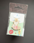A Peter Rabbit enamel pin in its original packaging, featuring a rabbit holding a pink teacup on a light green backing card with a brown hanging tab, set against a dark gray background.