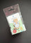 A Peter Rabbit enamel pin of Ned Flanders the cat, shown in its original clear plastic blister packaging with a white backing card. The pin depicts Ned wearing his signature blue apron.