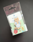 A Peter Rabbit enamel pin of Ned Flanders the cat, shown in its original clear plastic blister packaging with a white backing card. The pin depicts Ned wearing his signature blue apron.