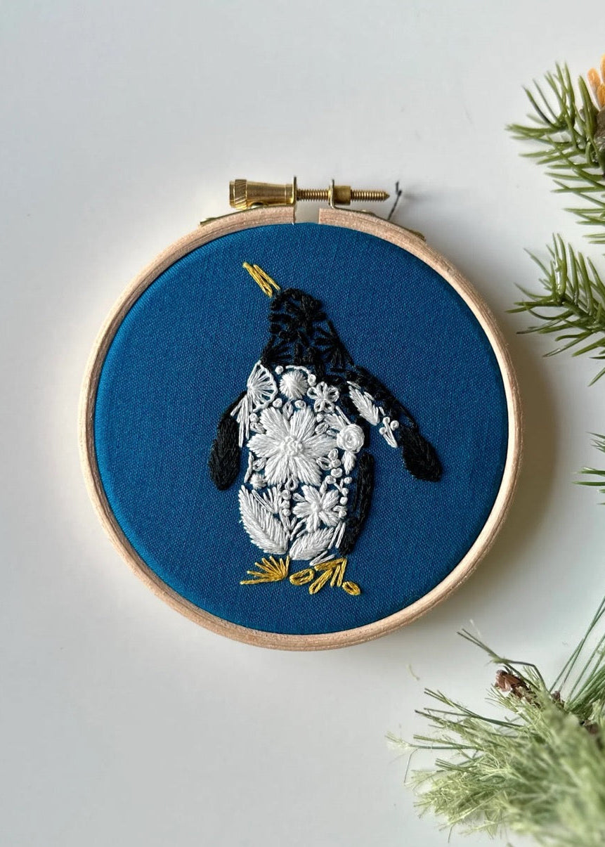 Embroidery hoop with a penguin design on blue fabric, surrounded by greenery.