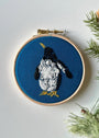 Embroidery hoop with a penguin design on blue fabric, surrounded by greenery.